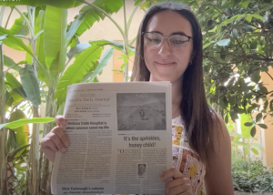 Morrow holds up a newspaper while standing among potted plants indoors.