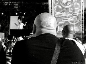 Rear view of a man seated at what appears to be a formal event or venue with stage lighting visible in the background.