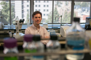 Professor Patrick Allard sits at a laboratory bench with scientific equipment and bottles visible in the foreground, with