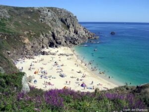 Porth Chapel beach in summer with golden sand, turquoise water, purple wildflowers on cliffs, and beachgoers enjoying the