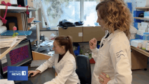 Two researchers work in a laboratory setting, one seated at a computer workstation and another standing nearby, surrounded