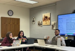 Three people sit at a table during a classroom discussion, with laptops and materials visible, while a presentation displays