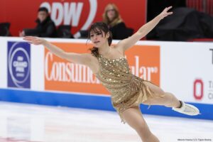 Alysa Liu performing a figure skating routine on ice, arms extended, wearing a gold sequined dress.