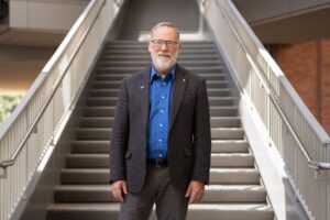 Fred Ramsdell stands on a staircase in a campus building, wearing a dark blazer and blue shirt.