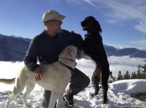 Ramsdell kneels in snow with two dogs during an outdoor winter scene with mountains in the background.
