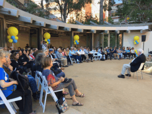 Outdoor memorial gathering with attendees seated in rows facing a speaker, with yellow balloons and trees in background.