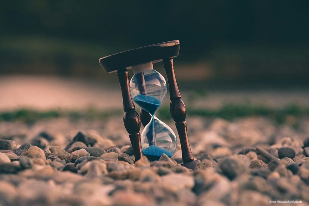 An hourglass with blue sand sits on a bed of small pebbles against a blurred background.