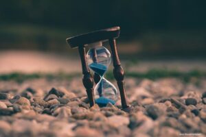 An hourglass with blue sand sits on a bed of small pebbles against a blurred background.
