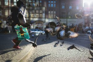 A person wearing red sneakers and carrying a colorful tote bag with a cat illustration runs through a sunlit cobblestone city square, scattering a flock of pigeons into flight. Several birds are captured mid-air with wings spread. (Credit: Itay Attias/UnSplash)