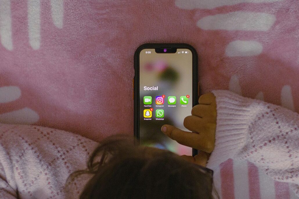 Child holding a smartphone displaying a social media folder with messaging and social apps against a pink background, suggesting mobile communication and social media use.