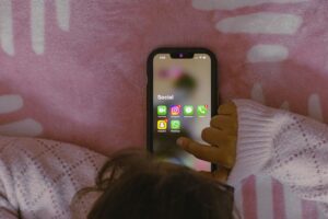 Child holding a smartphone displaying a social media folder with messaging and social apps against a pink background, suggesting mobile communication and social media use.